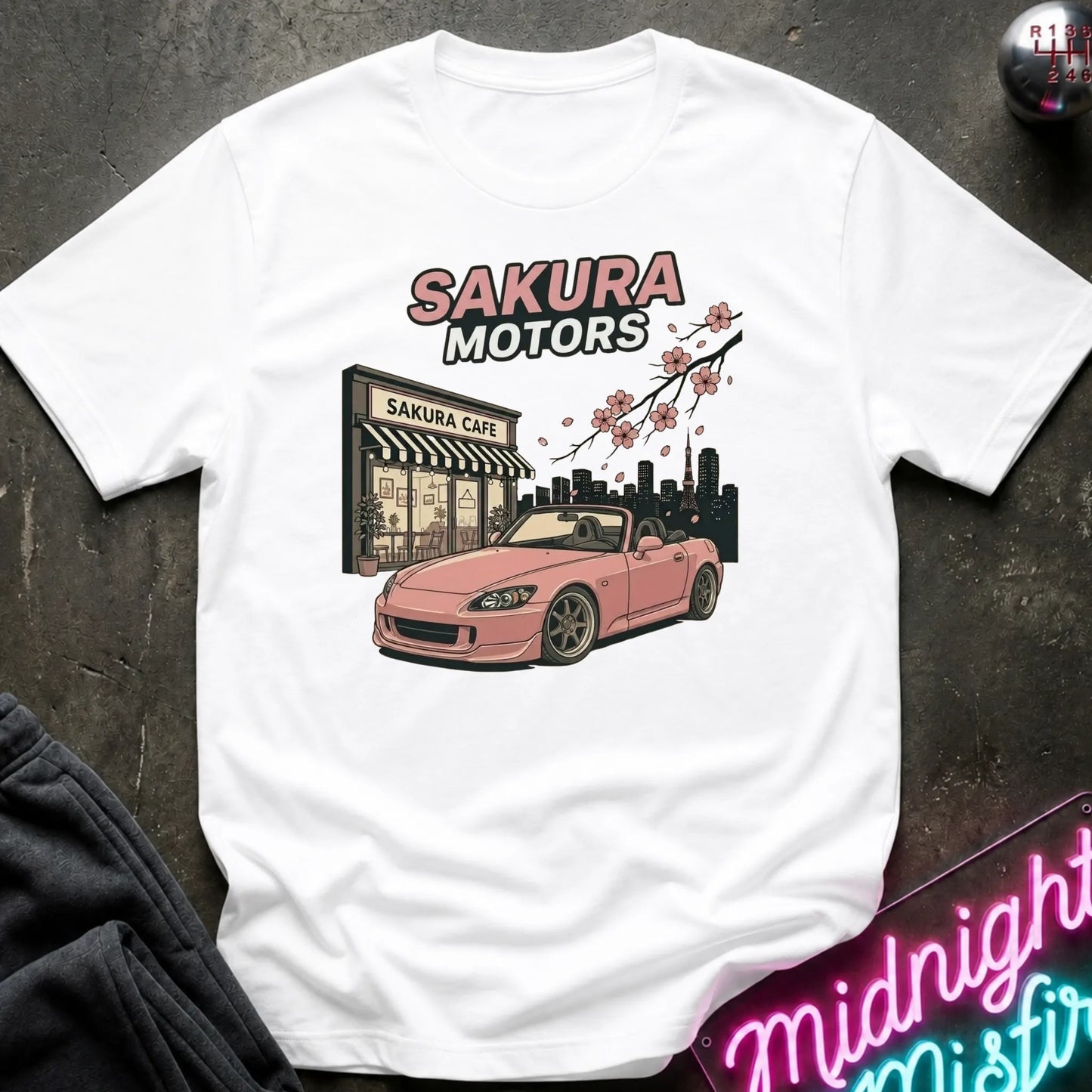 A white short-sleeve graphic T-shirt laid flat on a dark textured surface. The shirt features a printed design of a pink convertible sports car in front of a building labeled 'Sakura Cafe', with 'SAKURA MOTORS' text above and cherry blossoms across the top.