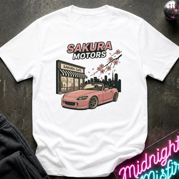 A white short-sleeve graphic T-shirt laid flat on a dark textured surface. The shirt features a printed design of a pink convertible sports car in front of a building labeled 'Sakura Cafe', with 'SAKURA MOTORS' text above and cherry blossoms across the top.