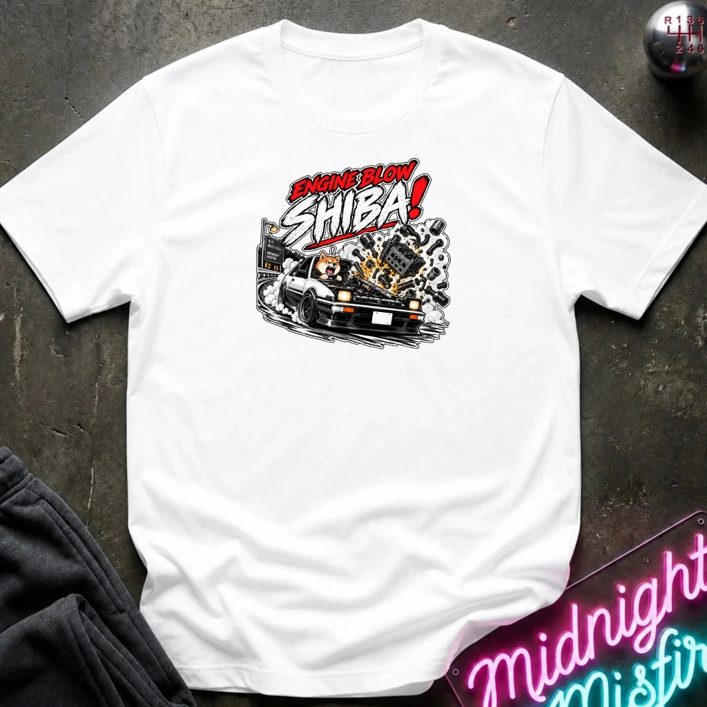 A white short-sleeve graphic t-shirt laid flat on a dark textured surface. The shirt features a stylized graphic of a white sports car with a glowing engine and the text 'ENGINE BLOW SHIBA!' in red, white, and black.