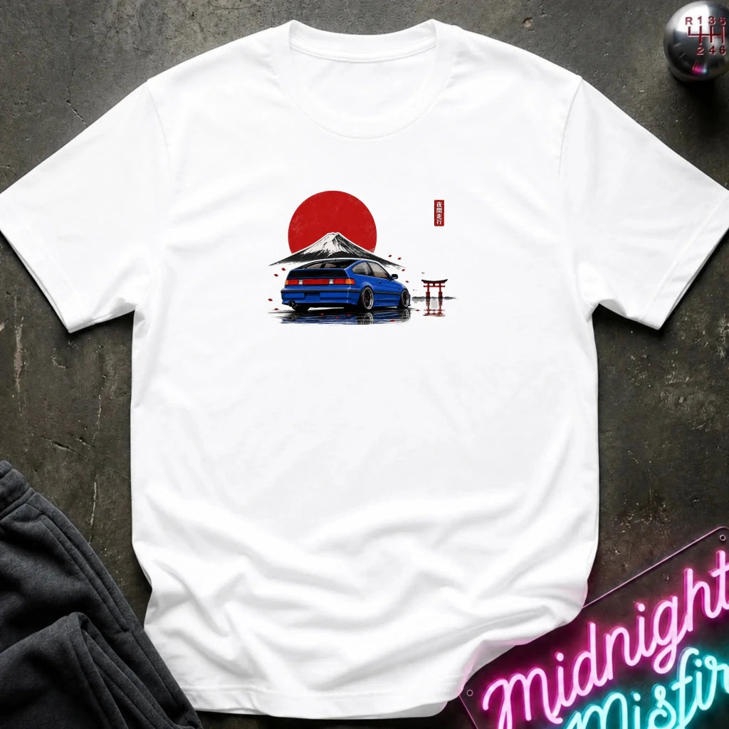 A white short-sleeve t-shirt laid flat on a dark textured surface, featuring a graphic print of a blue car in front of a red sun and Mount Fuji with a small red torii gate.