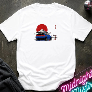A white short-sleeve t-shirt laid flat on a dark textured surface, featuring a graphic print of a blue car in front of a red sun and Mount Fuji with a small red torii gate.