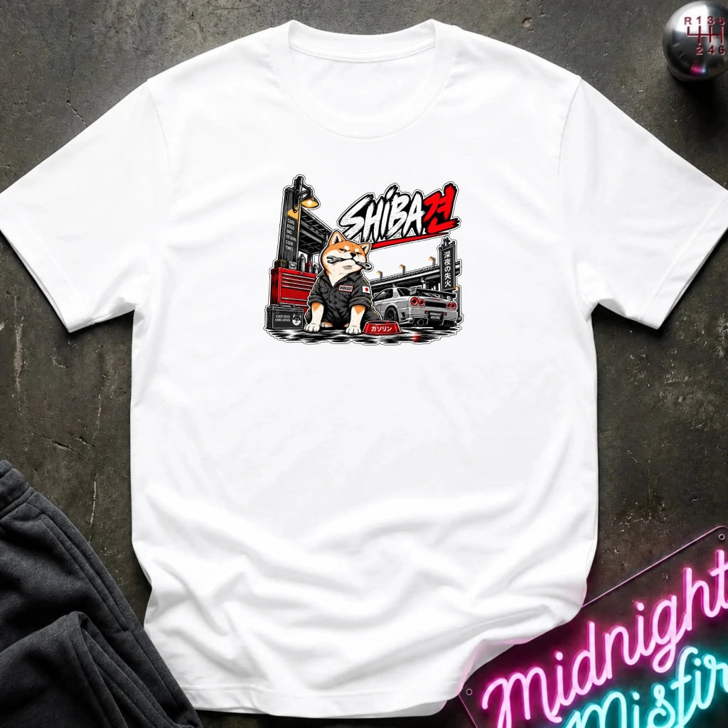 A white short-sleeve t-shirt laid flat on a dark textured surface, featuring a graphic print of a bulldog in a driver's helmet behind a race car with the text 'SHIBA'.