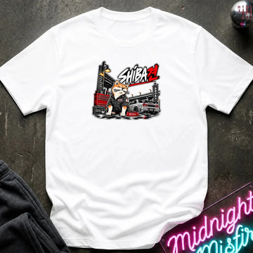 A white short-sleeve t-shirt laid flat on a dark textured surface, featuring a graphic print of a bulldog in a driver's helmet behind a race car with the text 'SHIBA'.