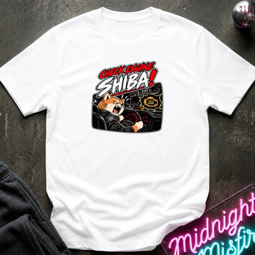 A white short-sleeve crewneck t-shirt laid flat on a dark textured surface. The shirt features a graphic print of an angry Shiba Inu dog in a racing car with the text 'CHECK ENGINE SHIBA!'.