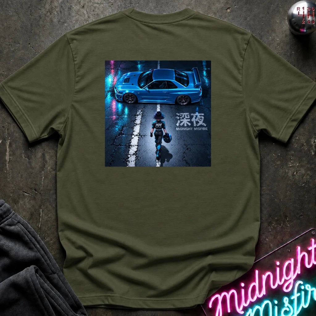 An olive green crewneck t-shirt laid flat on a dark textured surface, featuring a large graphic print on the chest of a blue sports car on a wet street at night with neon reflections and a figure walking in the middle.