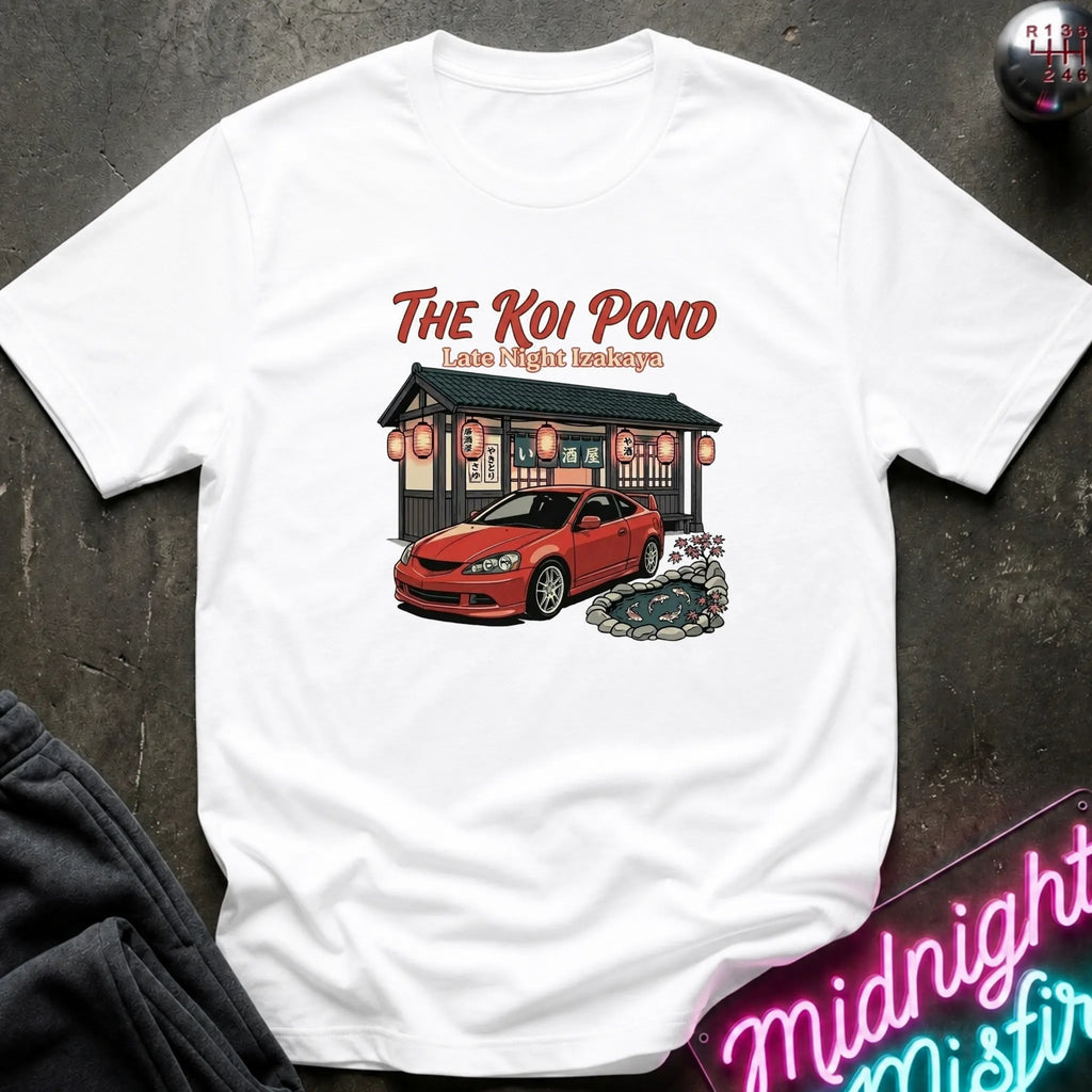 A white short-sleeve graphic t-shirt laid flat on a dark textured surface. The graphic features the text 'THE KOI POND Late Night Hamburger' above an illustration of a red sports car parked in front of a restaurant with red lanterns and a koi pond.