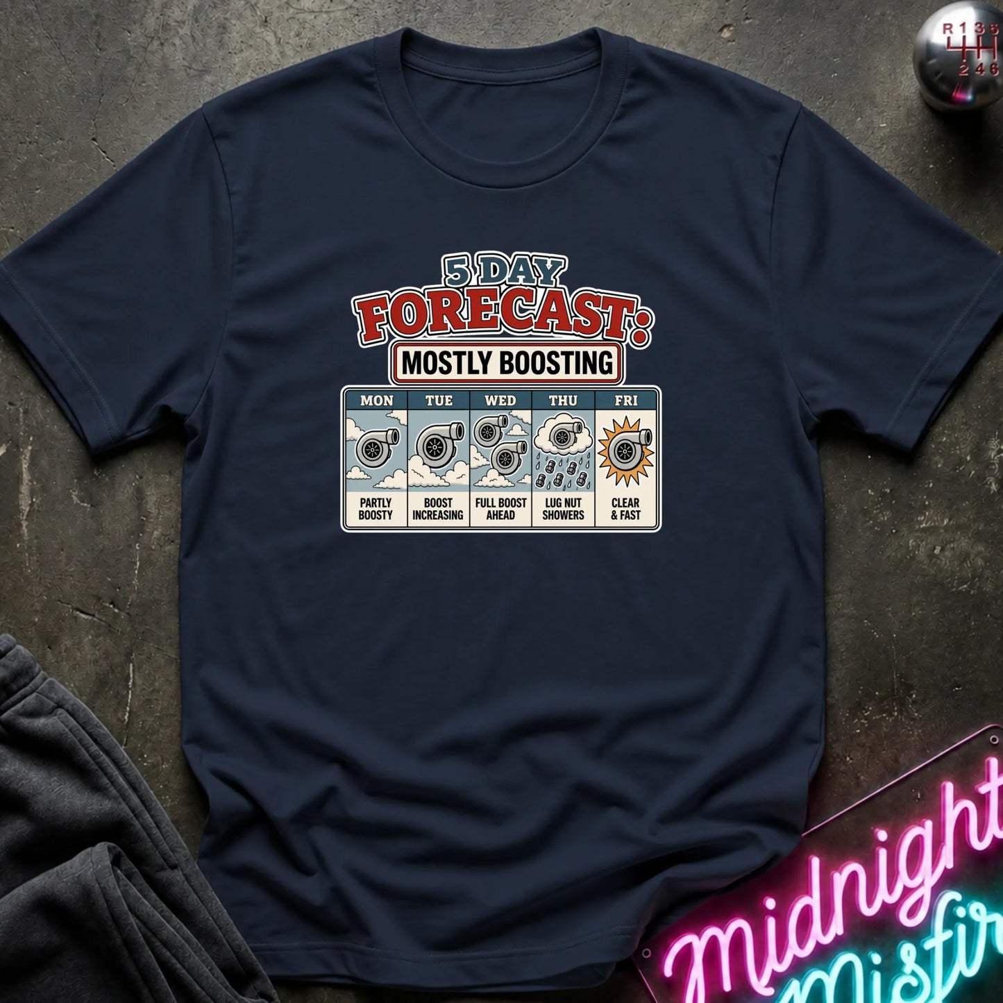 A navy blue short-sleeve graphic T-shirt laid flat on a dark textured surface. The shirt features a colorful '3 Day Forecast: Mostly Boosting' print with weather icons and text for Monday through Friday.