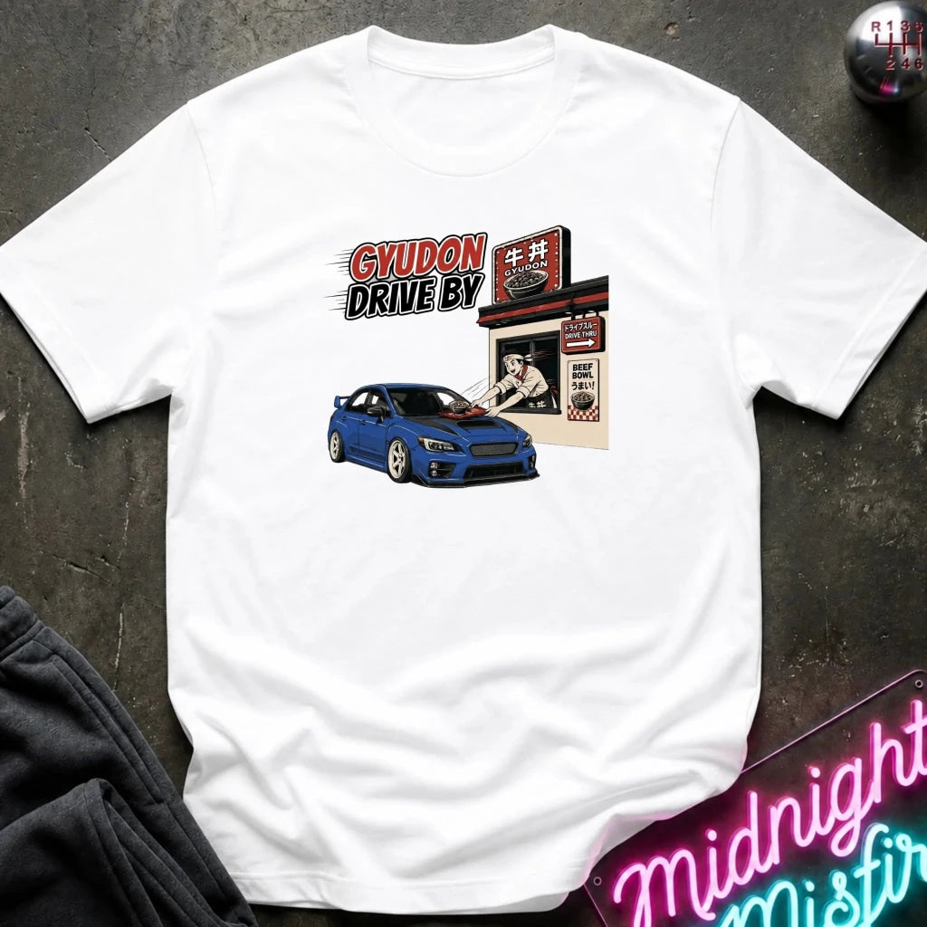 A white short-sleeve graphic t-shirt laid flat on a dark textured surface. The graphic features a blue sports car and a retro-style diner sign with the text 'GYUDON DRIVE BY'.