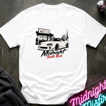 A white short-sleeve graphic T-shirt laid flat on a dark textured surface. The shirt features a black and white illustration of a sports car parked outside a cafe, with the text 'Midnight Taste Run' in black and red script below.