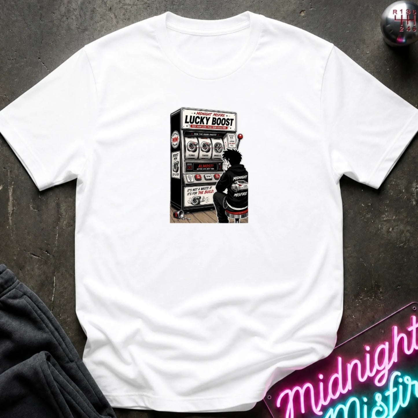 A white short-sleeve crewneck t-shirt laid flat on a dark textured surface, featuring a central graphic of a person sitting in front of a slot machine labeled 'LUCKY BOOST'.