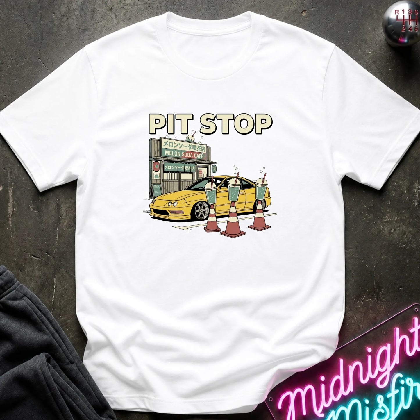 A white short-sleeve crewneck t-shirt featuring a cartoon graphic of a yellow car at a 'Pit Stop' gas station, styled flat on a dark textured background with a neon sign and dark pants partially visible.