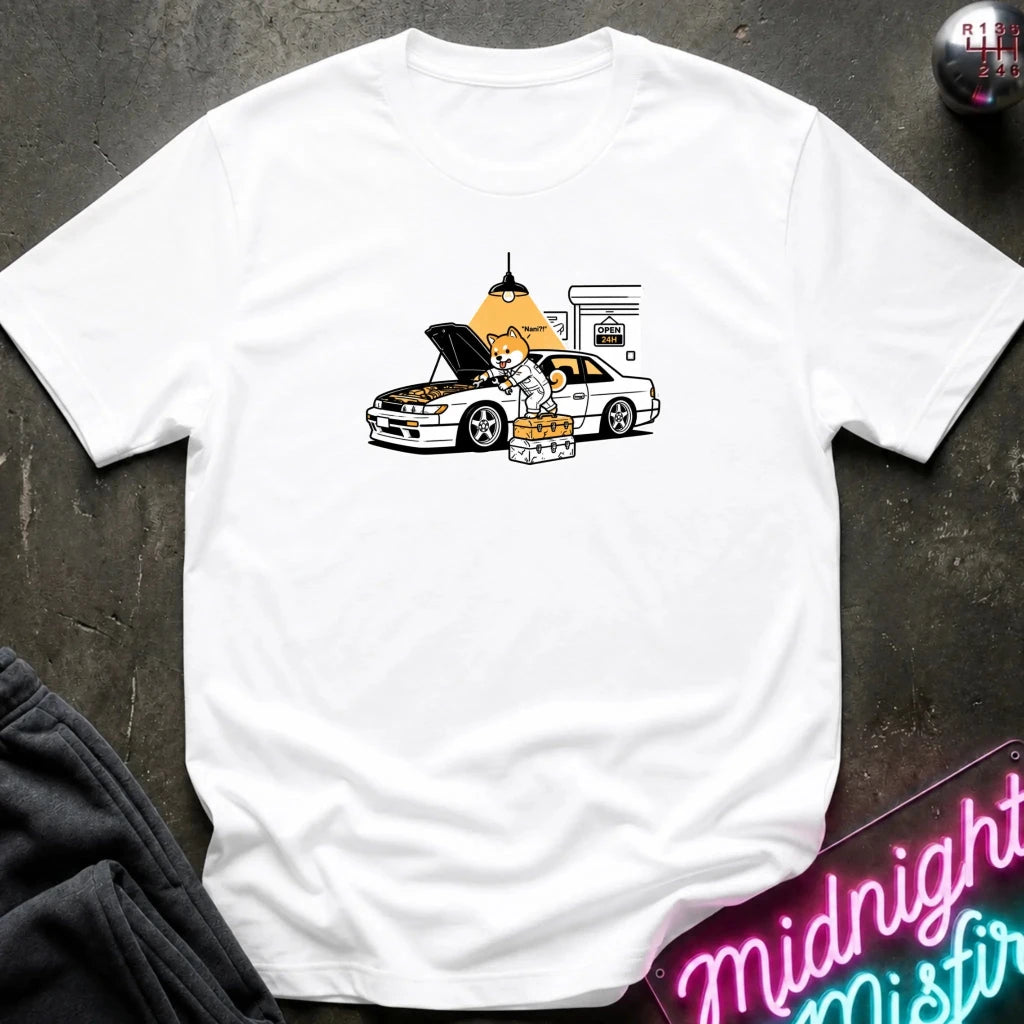 white graphic t-shirt laid flat on a dark textured surface. The graphic features a dog fixing a car.