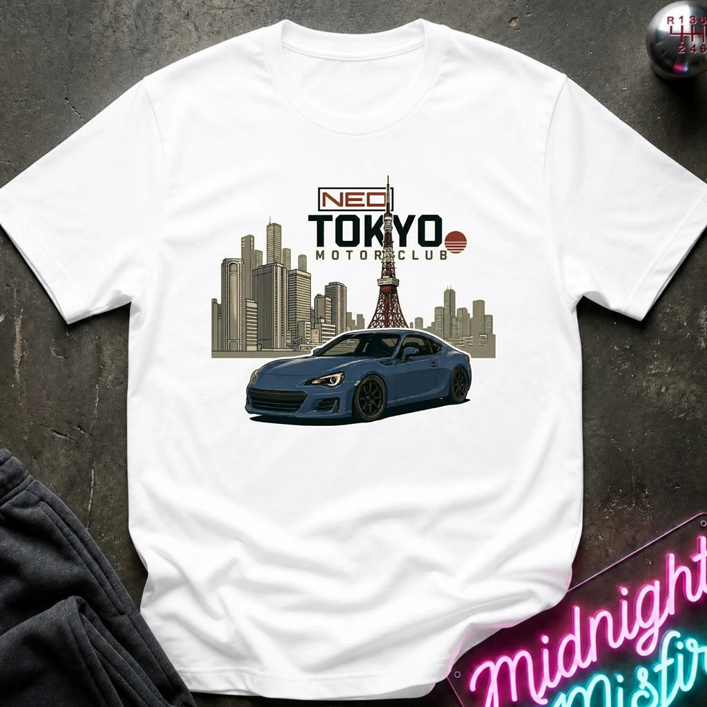 A white short-sleeve graphic T-shirt laid flat on a dark textured surface. The shirt features a printed design of a blue sports car in front of a Tokyo city skyline with the Tokyo Tower, and text reading 'NEO TOKYO MOTORCLUB'.