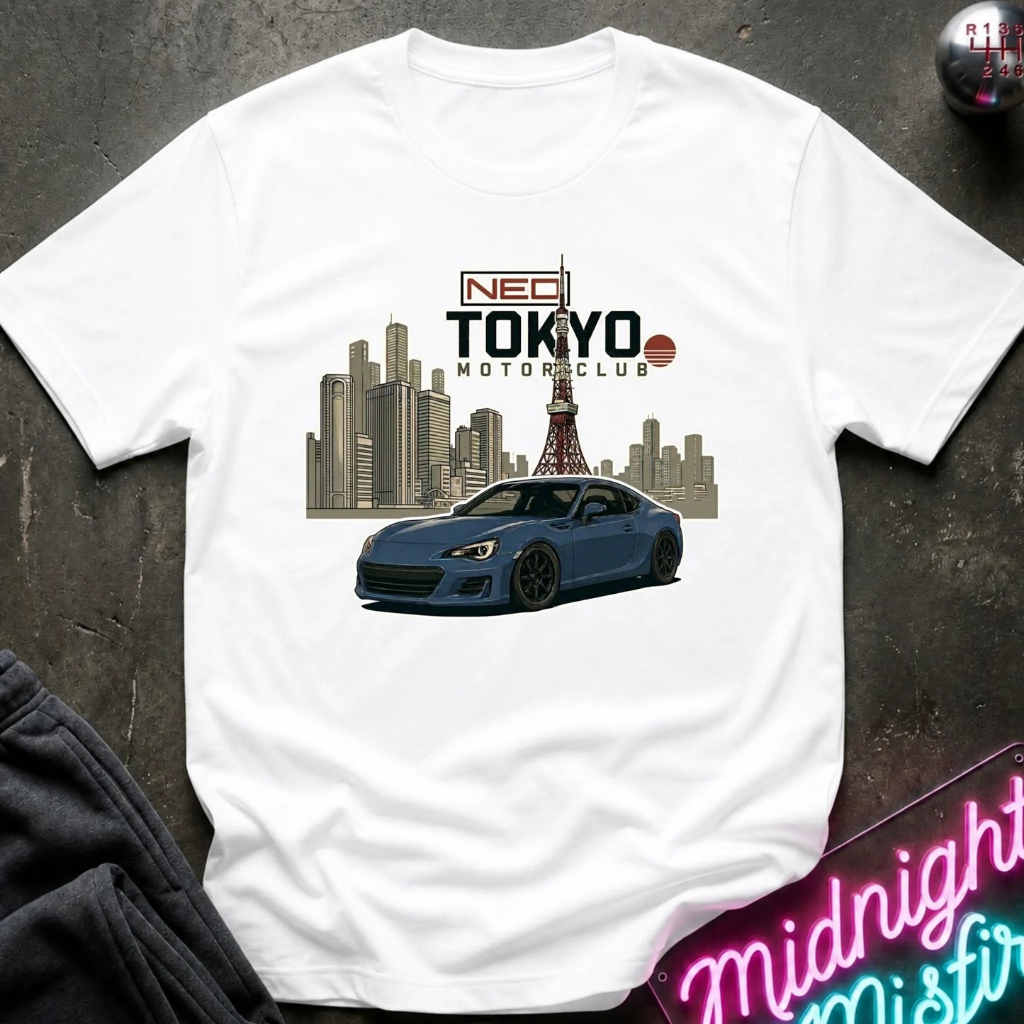 A white short-sleeve graphic T-shirt laid flat on a dark textured surface. The shirt features a printed design of a blue sports car in front of a Tokyo city skyline with the Tokyo Tower, and text reading 'NEO TOKYO MOTORCLUB'.