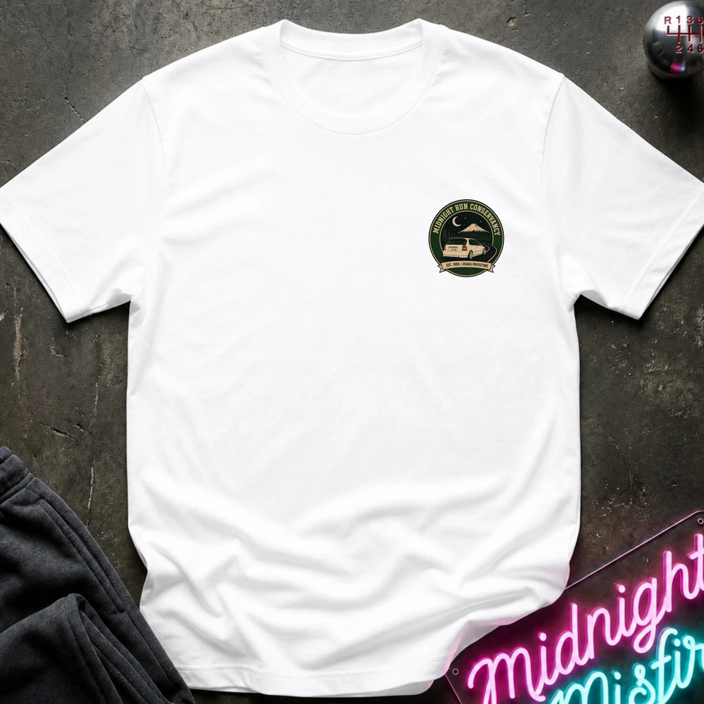 A white short-sleeve crewneck t-shirt with a circular green and black chest graphic, styled flat-lay on a dark textured surface with a neon sign and dark pants nearby.