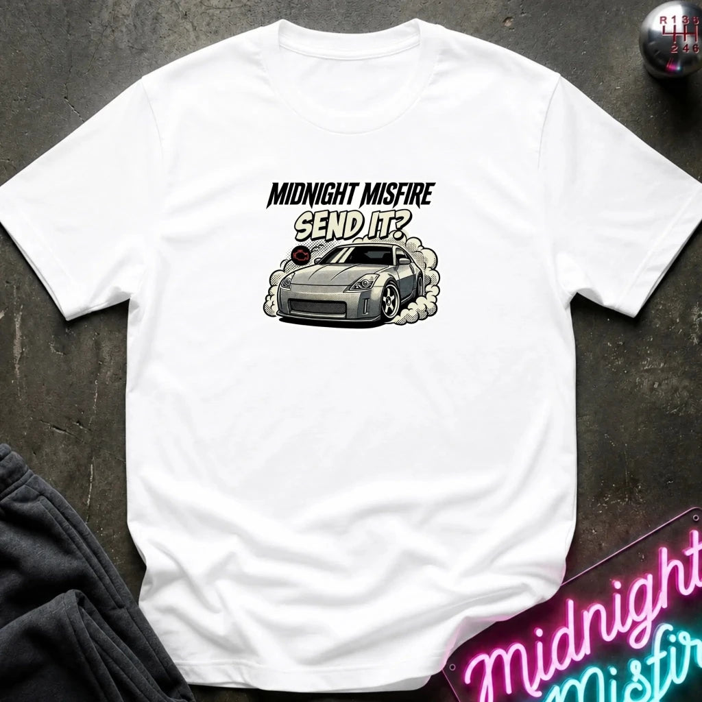 A white short-sleeve t-shirt laid flat on a dark textured surface, featuring a graphic of a gray sports car with smoke and the text 'MIDNIGHT MISFIRE SEND IT?' printed on the chest.