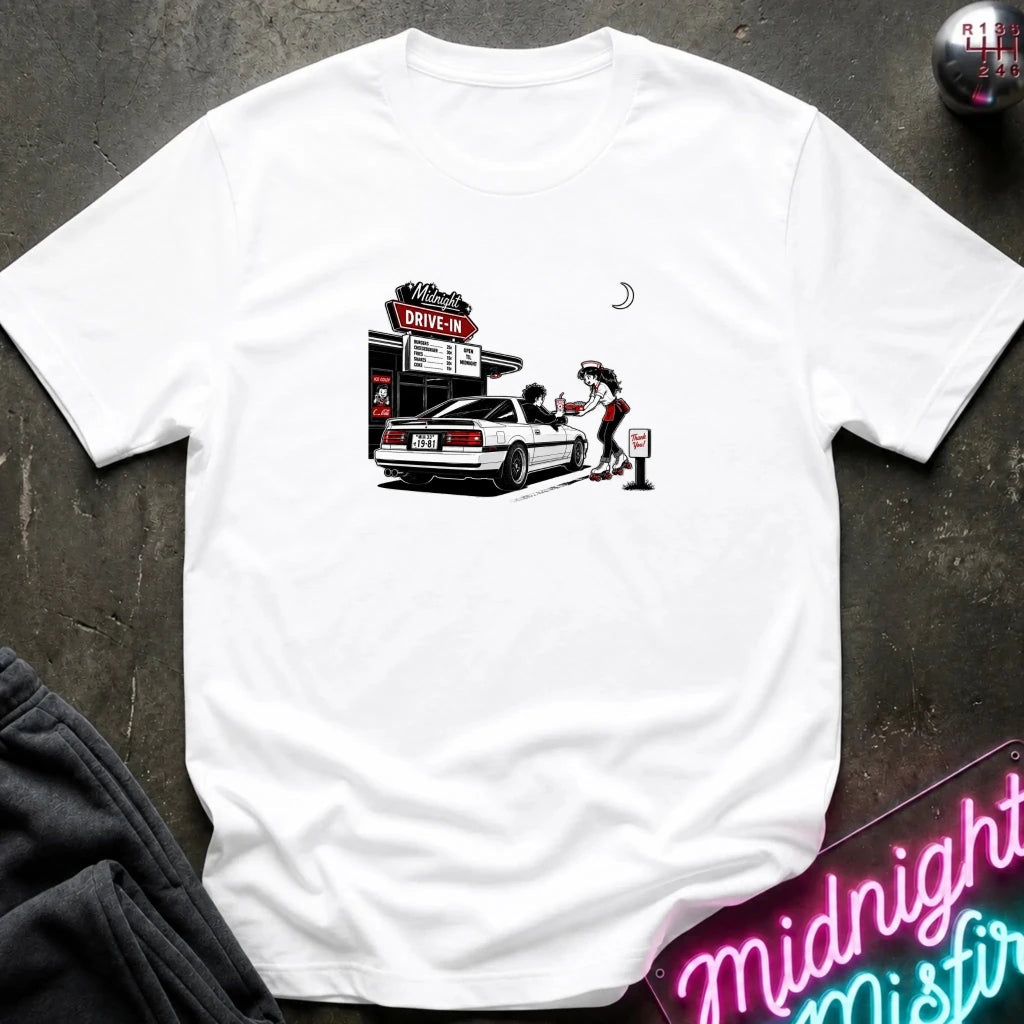 A white short-sleeve t-shirt laid flat on a dark textured surface, featuring a graphic print of a woman pumping gas at a drive-in station next to a white car. 