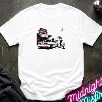 A white short-sleeve t-shirt laid flat on a dark textured surface, featuring a graphic print of a woman pumping gas at a drive-in station next to a white car. 