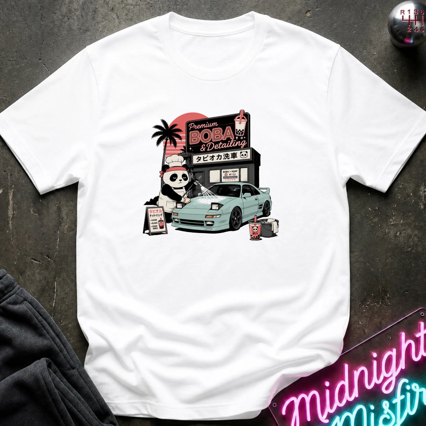 A white short-sleeve graphic T-shirt laid flat on a dark textured surface. The shirt features a colorful retro-style print of a panda character, a light blue sports car, and a sign reading 'Premium BOBA & Coffee' with Japanese text.