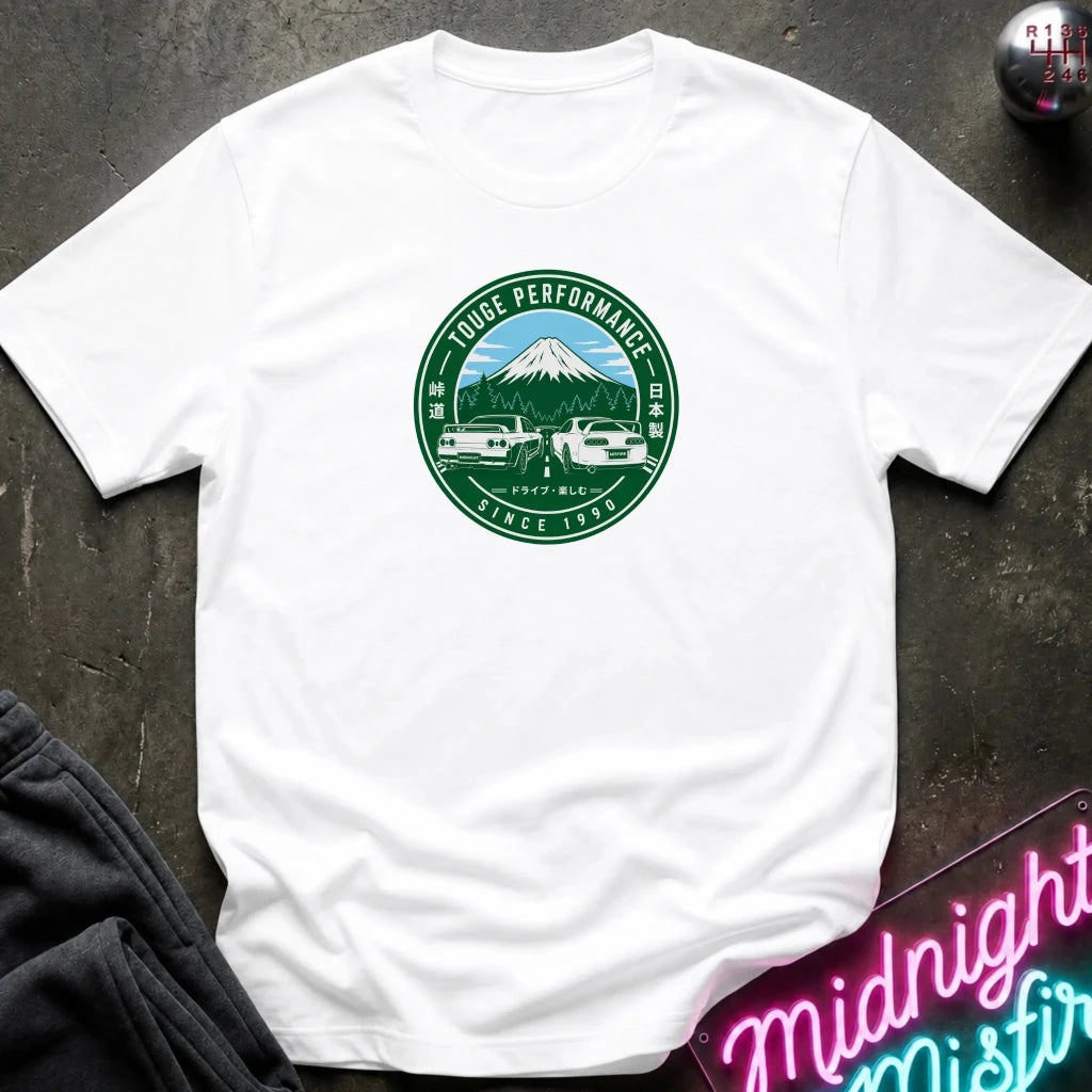 A white short-sleeve crewneck t-shirt laid flat on a dark textured surface. The shirt features a circular green and blue graphic on the chest depicting a mountain, two cars, and the text 