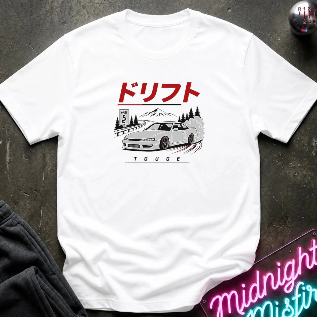 A white short-sleeve graphic T-shirt laid flat on a dark textured surface. The shirt features a printed illustration of a white sports car with a red sun and mountain graphic above it, along with small red and black text.