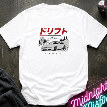 A white short-sleeve graphic T-shirt laid flat on a dark textured surface. The shirt features a printed illustration of a white sports car with a red sun and mountain graphic above it, along with small red and black text.