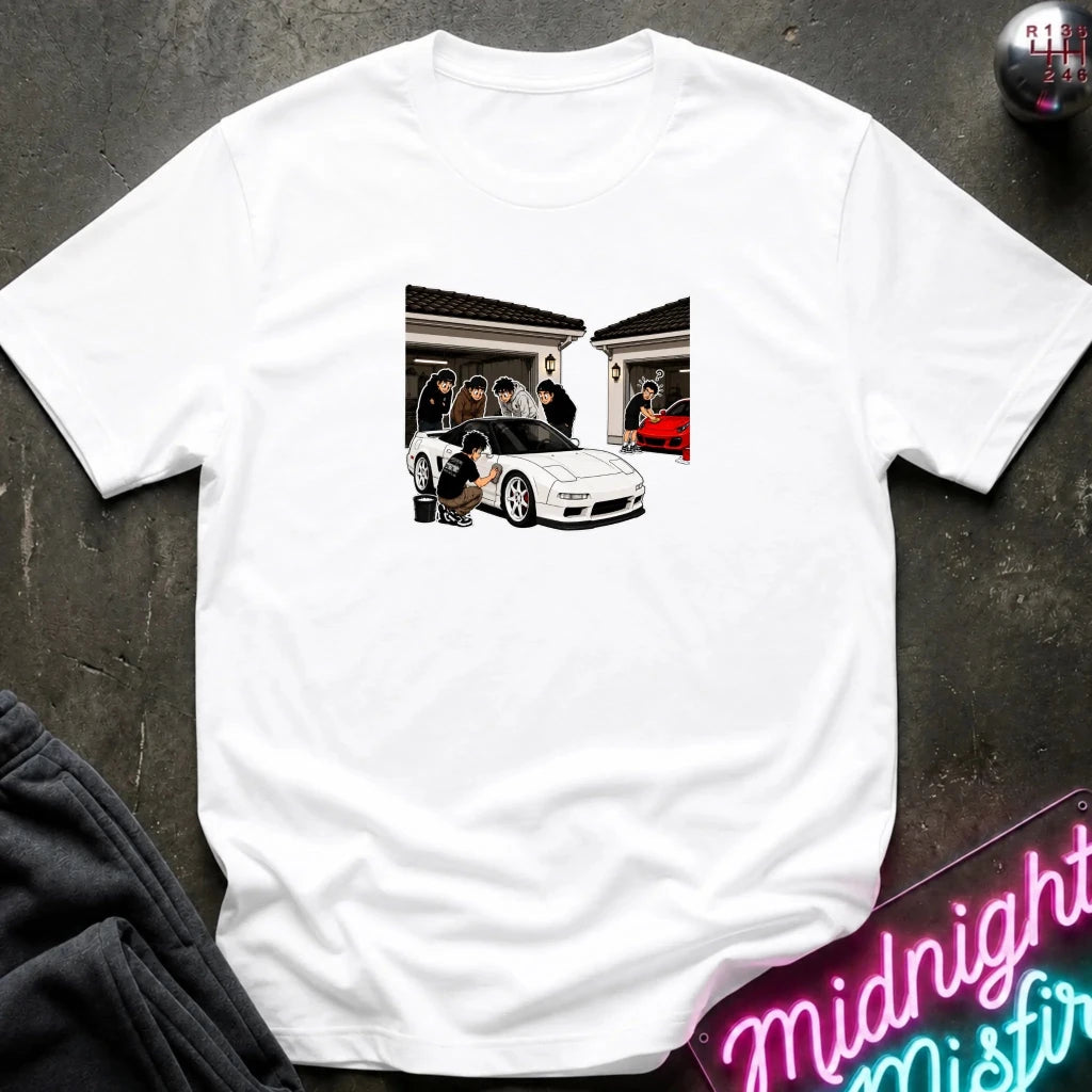 A white short-sleeve crewneck t-shirt laid flat on a dark textured surface. The center of the shirt features a rectangular graphic print depicting a white sports car in front of a house with people standing by the garage.