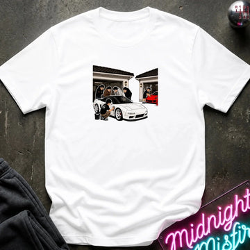 A white short-sleeve crewneck t-shirt laid flat on a dark textured surface. The center of the shirt features a rectangular graphic print depicting a white sports car in front of a house with people standing by the garage.