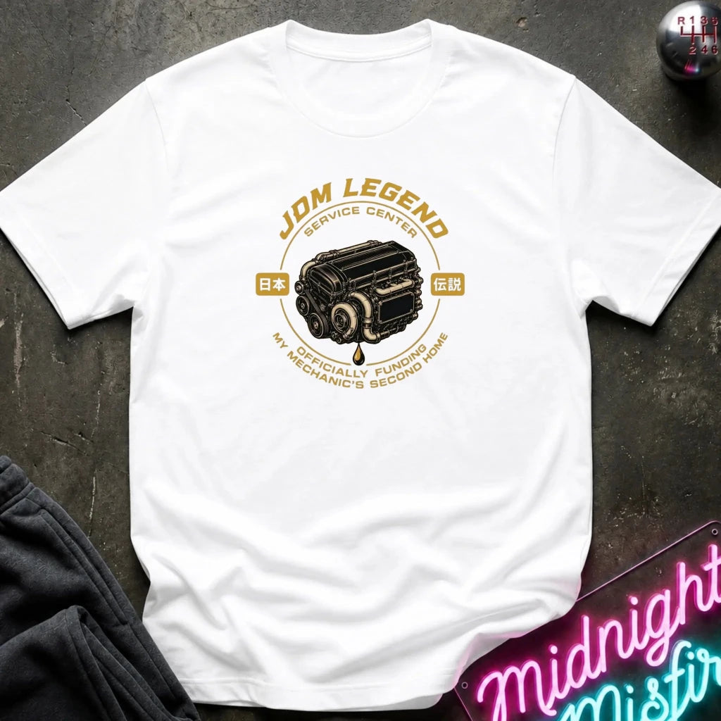 white graphic t-shirt laid flat on a dark textured surface. The shirt features a vintage-style yellow and black engine graphic with the text 'JDM LEGEND' and 'MY SPECIALTY FUNDS MECHANIC'S SECOND HAND'.