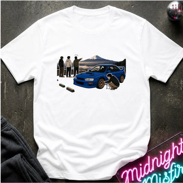 A white short-sleeve crewneck t-shirt laid flat on a dark textured surface. The shirt features a graphic print of a blue sports car with a black spoiler, a mountain background, and three people standing nearby.