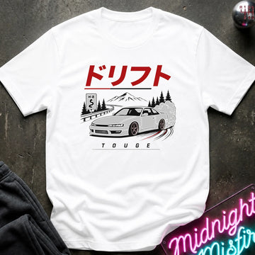 white  crewneck t-shirt featuring a graphic print of a white drift car on a road with mountains in the background, under red Japanese text. The shirt is laid flat on a dark textured surface with a neon sign and dark clothing partially visible in the corners.
