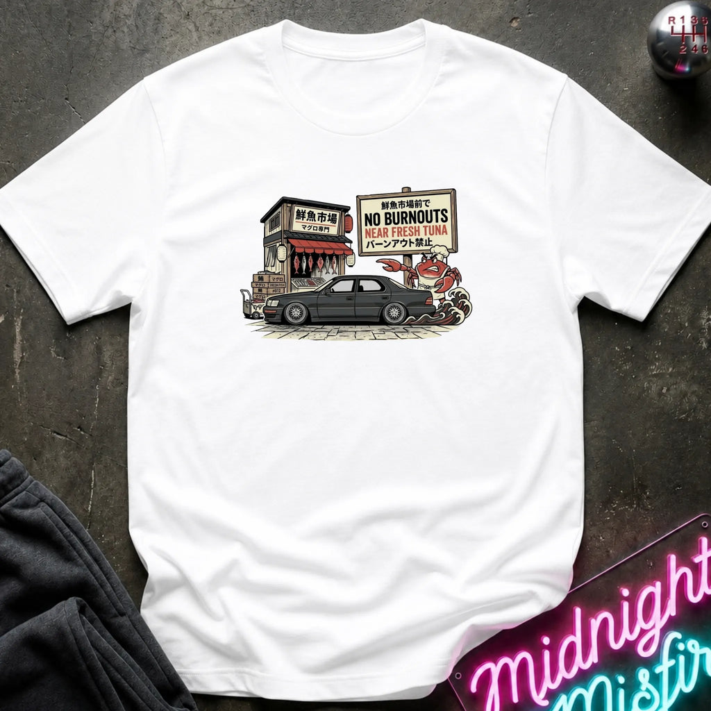 A white short-sleeve graphic t-shirt laid flat on a dark textured surface. The graphic shows a black car parked in front of a small building with a sign.