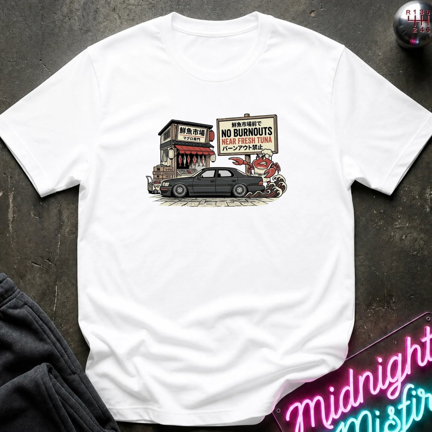 A white short-sleeve graphic t-shirt laid flat on a dark textured surface. The graphic shows a black car parked in front of a small building with a sign.