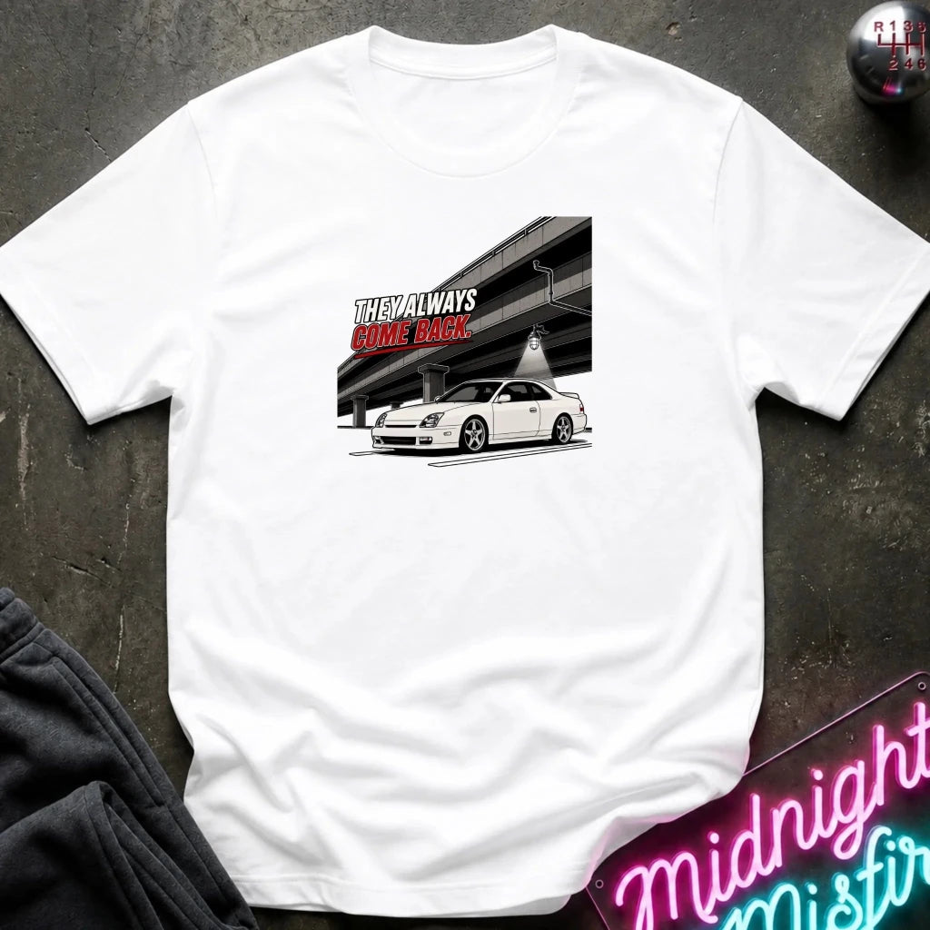 A white short-sleeve t-shirt laid flat on a dark textured surface, featuring a graphic print of a white sports car in front of an elevated highway 