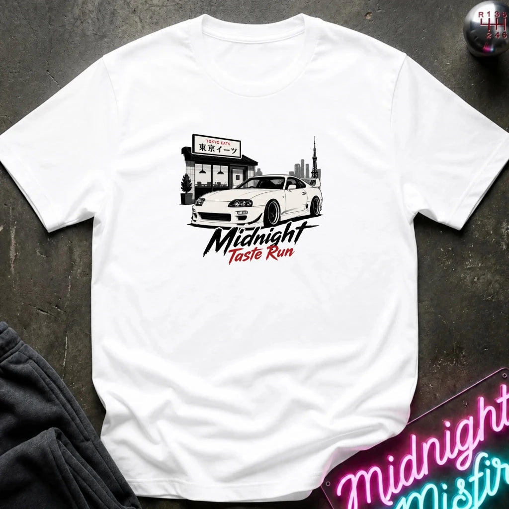A white short-sleeve graphic t-shirt laid flat on a dark textured surface. The graphic features a white sports car in front of a building with Japanese text, and the words 'Midnight Taste Run' in black and red script.