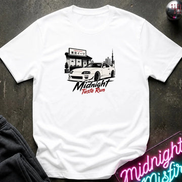 A white short-sleeve graphic t-shirt laid flat on a dark textured surface. The graphic features a white sports car in front of a building with Japanese text, and the words 'Midnight Taste Run' in black and red script.