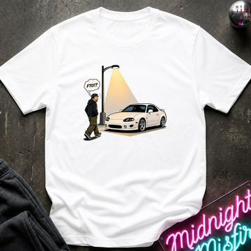 A white short-sleeve graphic t-shirt laid flat on a dark textured surface. The graphic shows a white sports car parked next to a streetlamp with a spotlight shining on a walking figure.