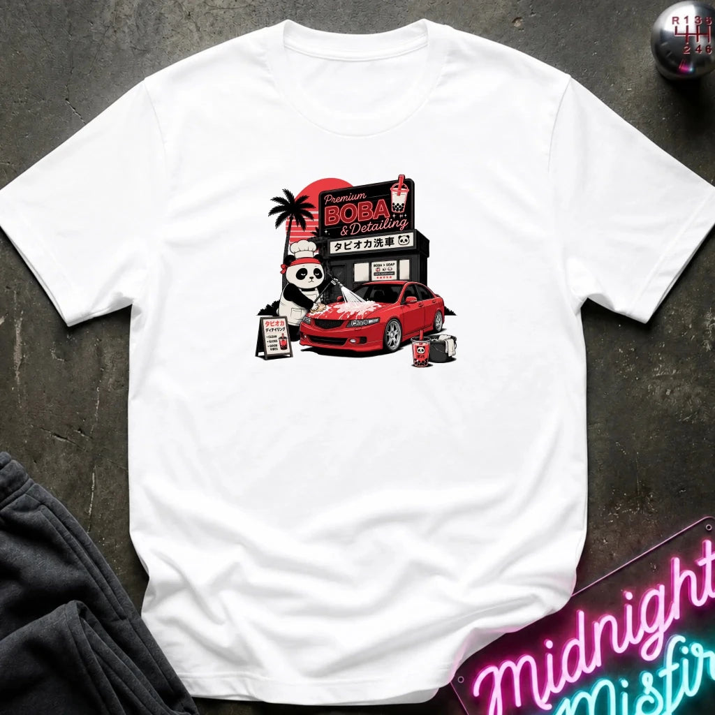 white short-sleeve graphic t-shirt laid flat on a dark textured surface. The graphic features a cartoon panda wearing a hat next to a red sports car, with a sign reading 'Midnight Boba'.