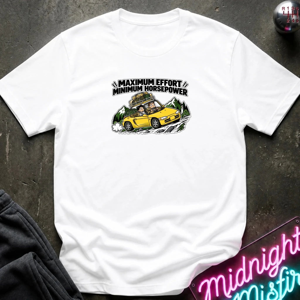 white graphic t-shirt laid flat on a dark textured surface. The graphic features a yellow sports car with a driver and passengers, with the text 'MAXIMUM EFFORT MINIMUM HORSEPOWER' above it.