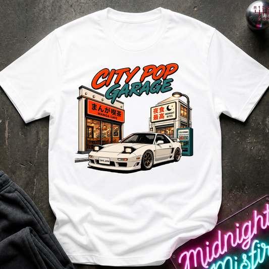 A white short-sleeve graphic T-shirt laid flat on a dark textured surface. The shirt features a colorful illustration of a white sports car parked outside a cafe with the text 'CITY POP GARAGE' above it.
