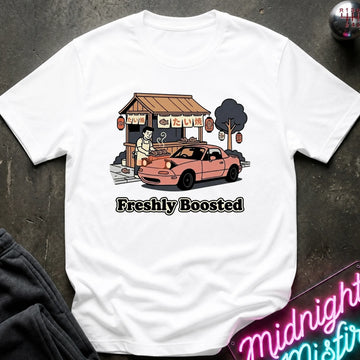 A white short-sleeve t-shirt laid flat on a dark textured surface, featuring a graphic of a pink sports car parked in front of a small food stall, with the text 'Freshly Boosted' printed below.