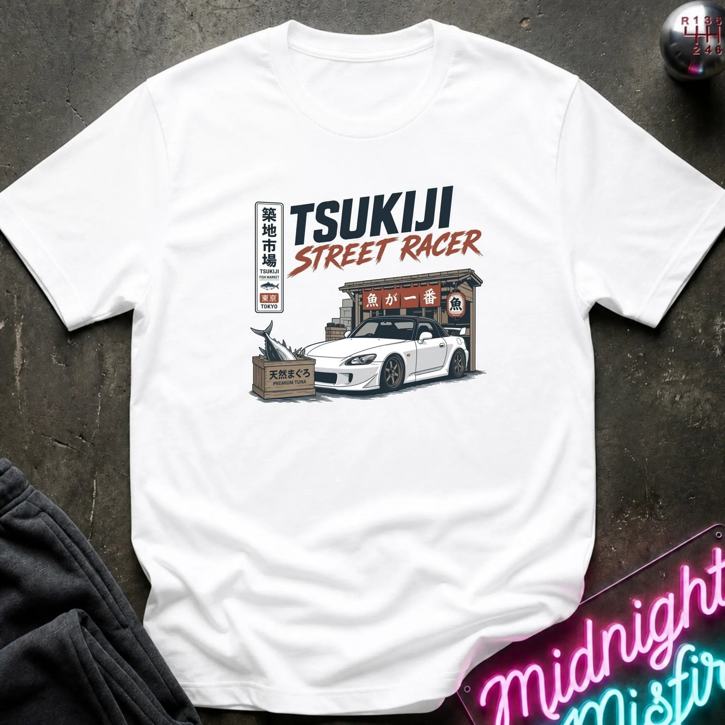 White short-sleeve graphic T-shirt laid flat on and surrounding an illustration of a white sports car parked outside a fish market.