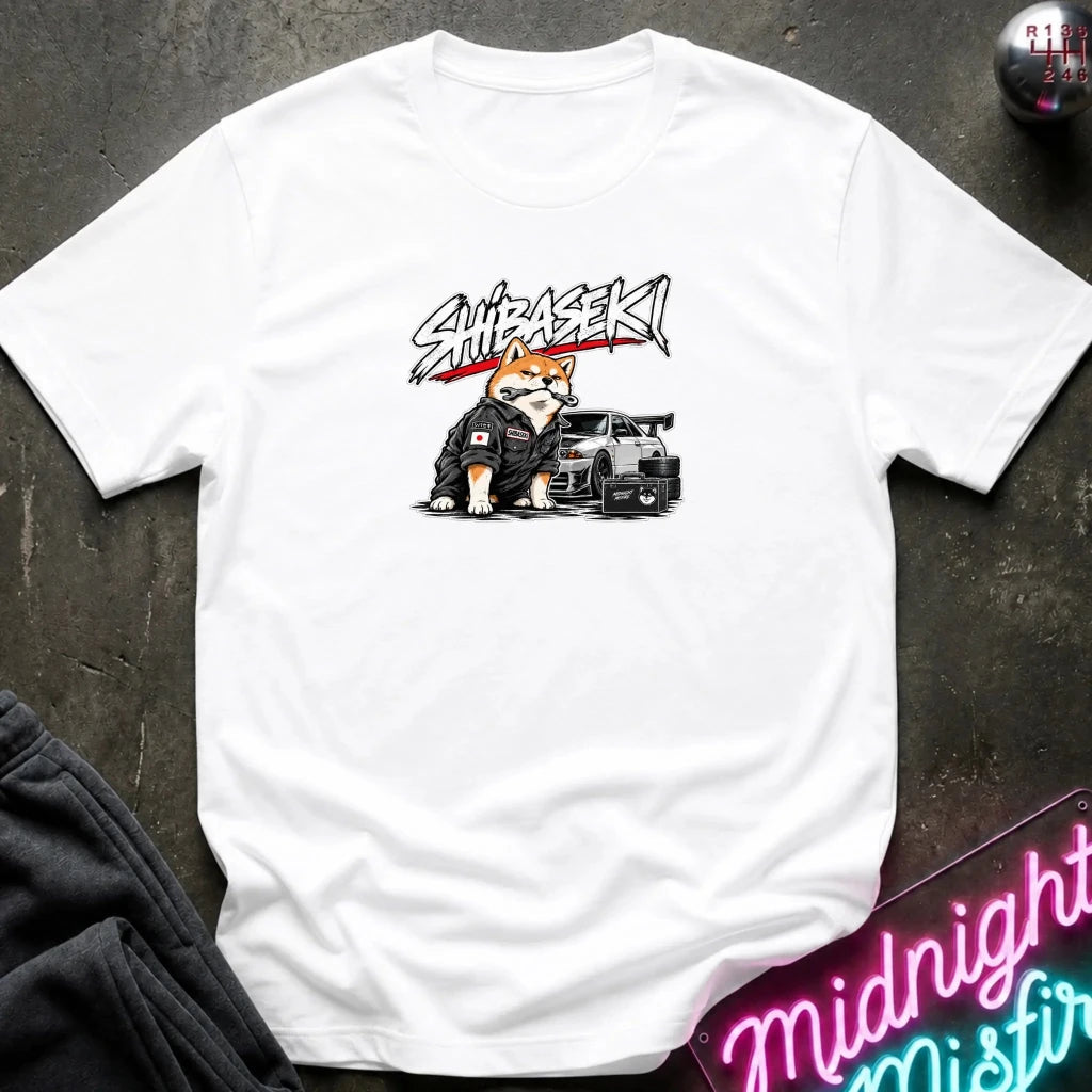 A white short-sleeve graphic t-shirt laid flat on a dark textured surface. The graphic features a Shiba Inu dog wearing a black racing suit next to a black sports car, with the word 'SHIBASEKI' written above in a stylized black and red font.