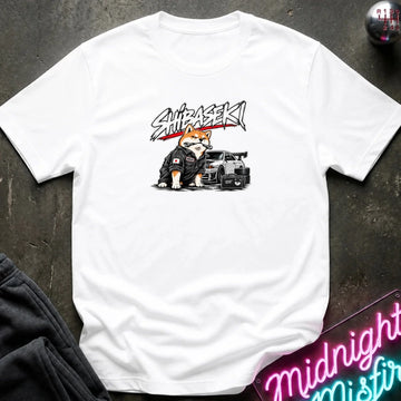 A white short-sleeve graphic t-shirt laid flat on a dark textured surface. The graphic features a Shiba Inu dog wearing a black racing suit next to a black sports car, with the word 'SHIBASEKI' written above in a stylized black and red font.