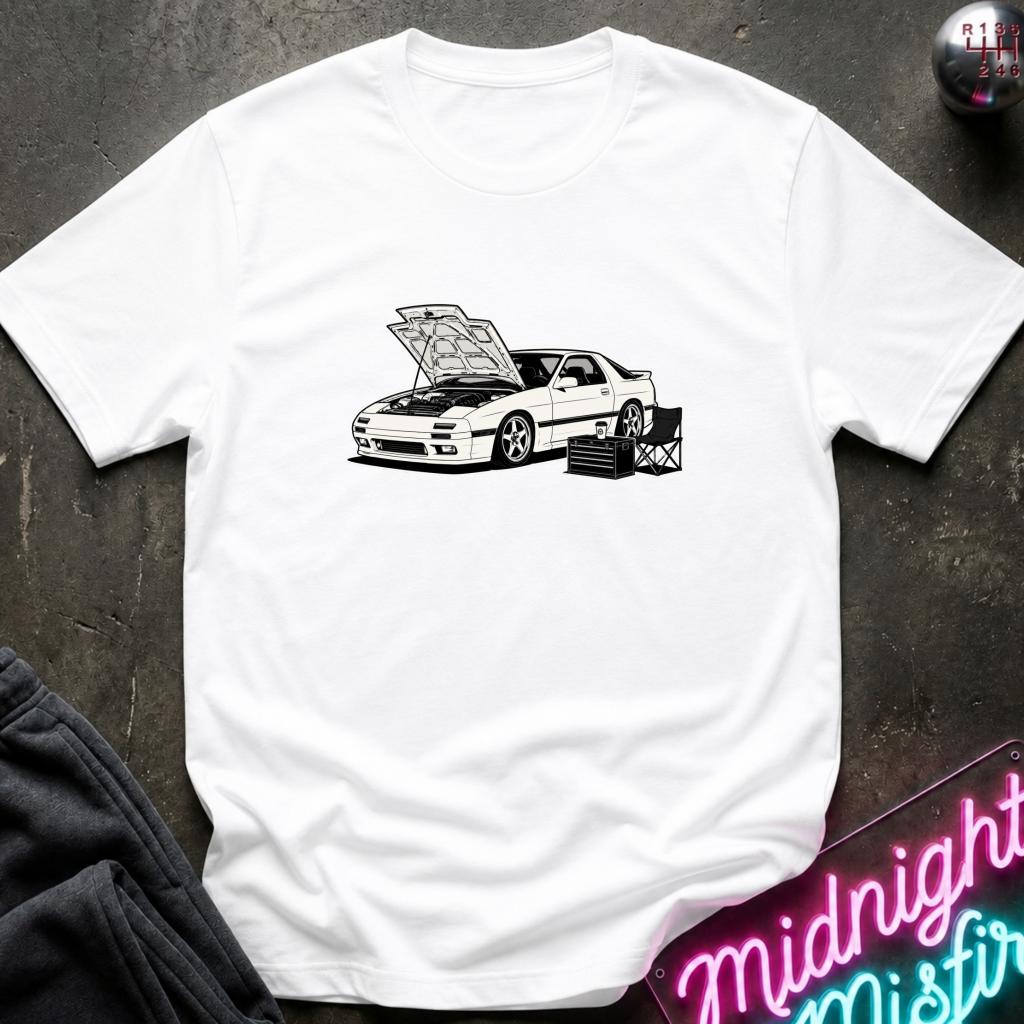 A white short-sleeve t-shirt laid flat on a dark textured surface, featuring a black line-art graphic of a sports car with its hood open and tool boxes underneath. A neon sign reading 'Midnight Misfire' is partially visible in the bottom right corner.