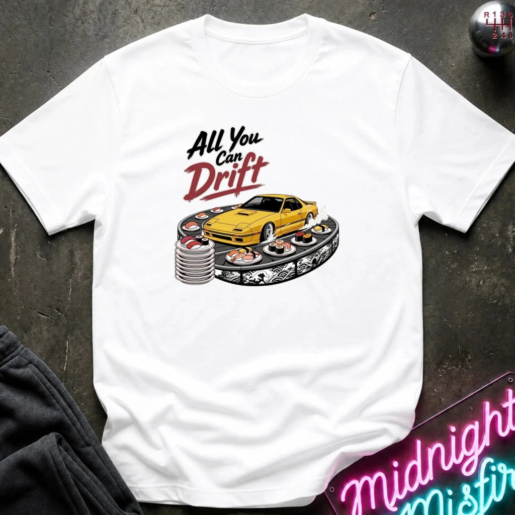 A white short-sleeve graphic T-shirt laid flat on a dark textured surface. The shirt features a printed design with the text 'All You Can Drift' above a yellow sports car positioned on stacked racing tires.