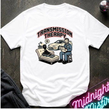 A white short-sleeve graphic t-shirt laid flat on a dark textured surface. The graphic features the text 'TRANSMISSION THERAPY' above an illustration of a therapist sitting on a stool with a patient in a chair, alongside a shifter.