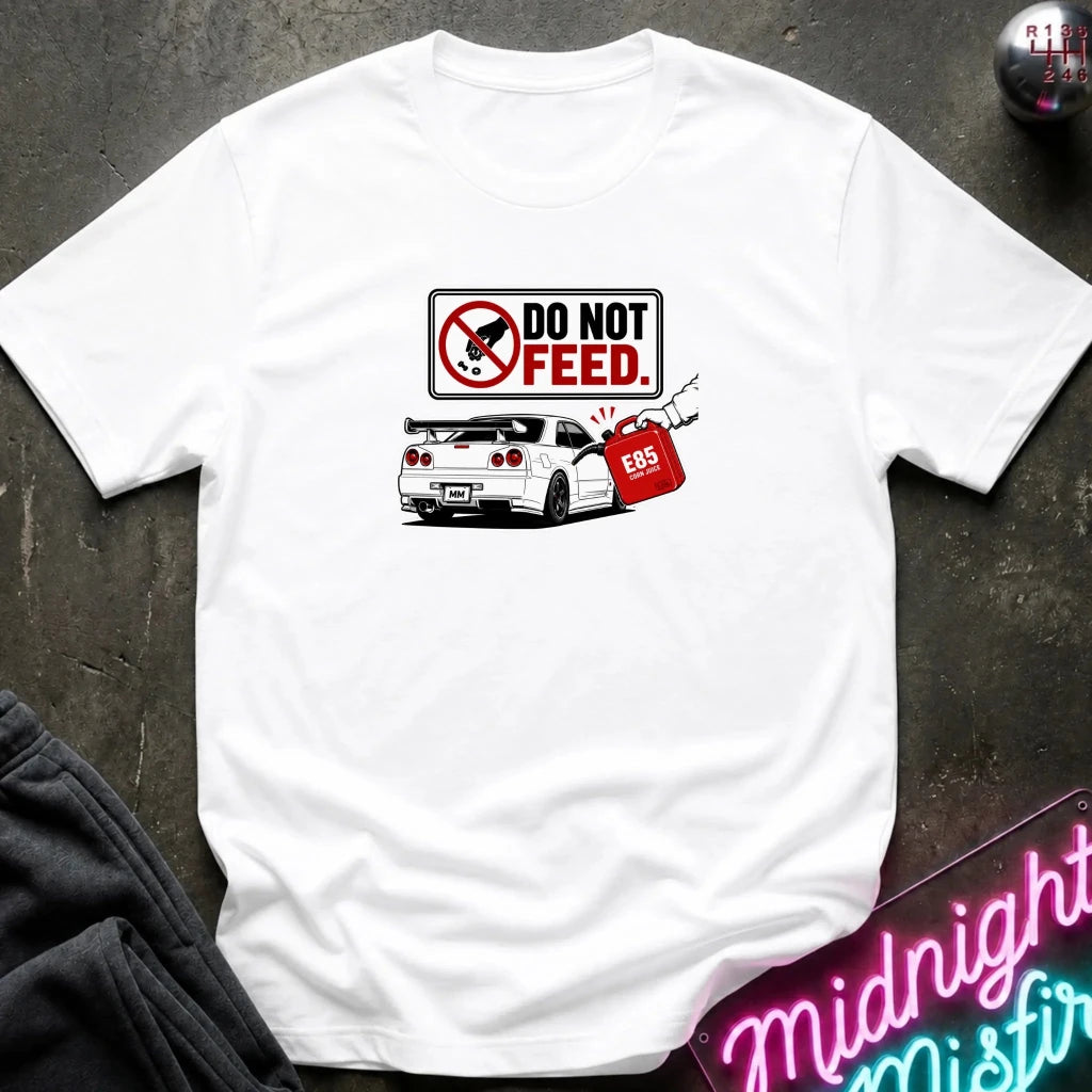 A white short-sleeve graphic t-shirt laid flat on a dark textured surface. The graphic features a 'DO NOT FEED' sign.
