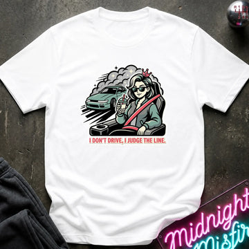 A white short-sleeve graphic t-shirt laid flat on a dark textured surface. The graphic shows a stylized woman in a car with a steering wheel, a green car, and the text 'I DON'T DRIVE, I JUDGE THE LINE.