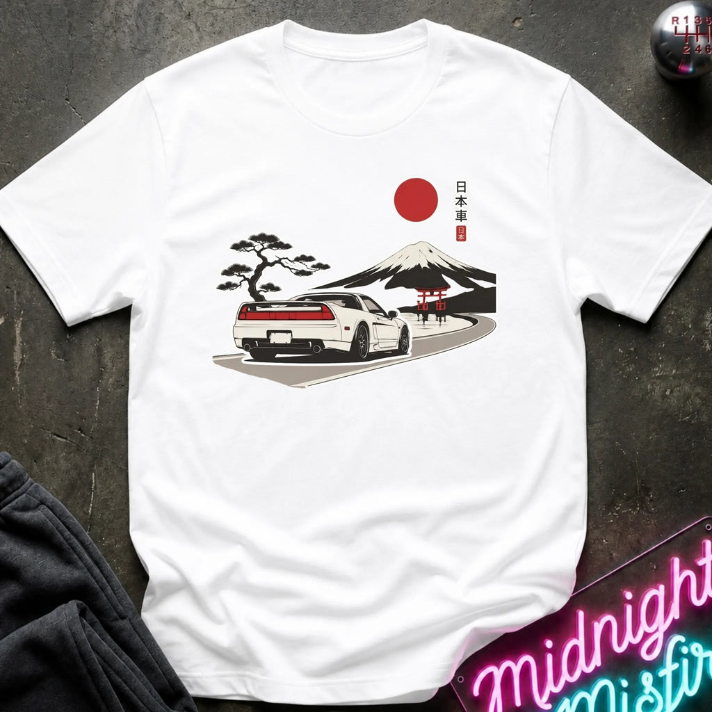 A white short-sleeve t-shirt laid flat on a dark textured surface, featuring a graphic print of a white sports car on a road with a red sun, Mount Fuji, a black pine tree, and a red torii gate, alongside Japanese text.